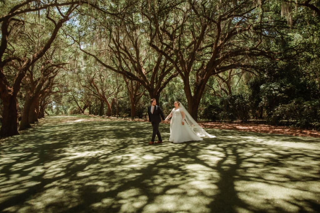 A wedding couple standing on a sunlit lawn beneath an oak canopy at Charles Towne Landing in Charleston.