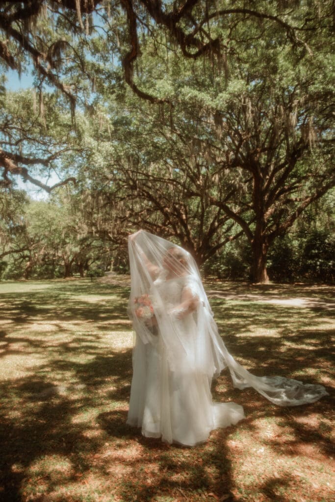 A bride holding a bouquet beneath her sheer veil, surrounded by oak trees and Spanish moss.