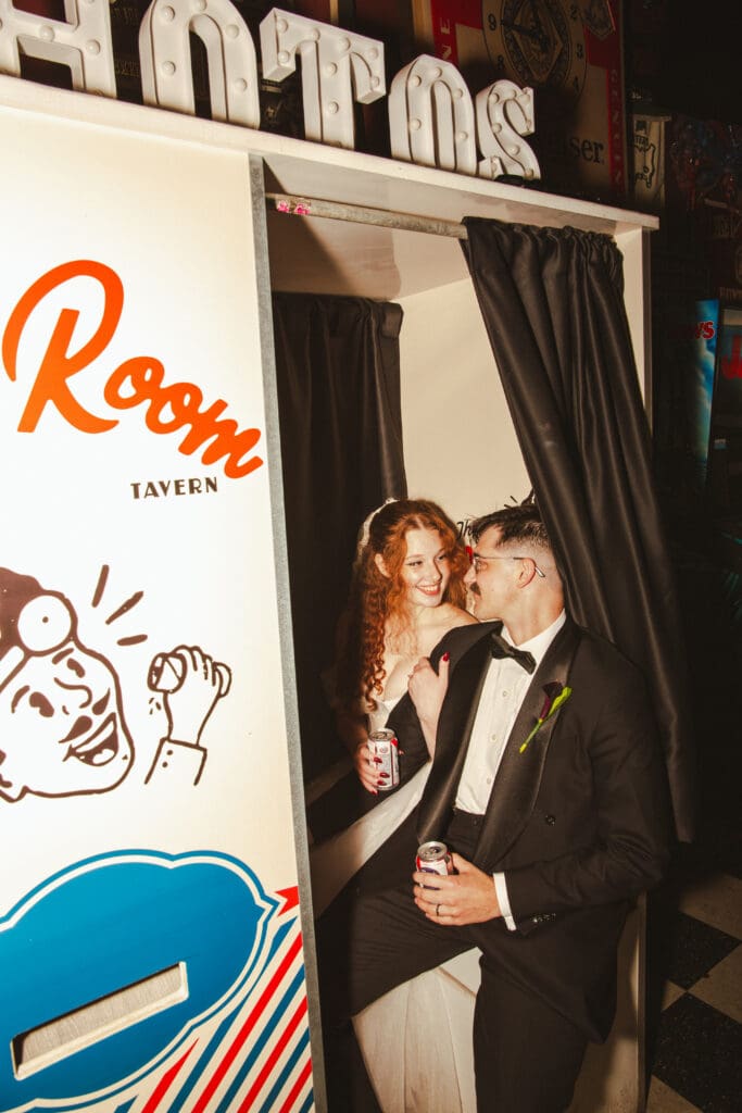 A newly married couple sharing a kiss and holding drinks in a photo booth at The Recovery Room in Charleston.