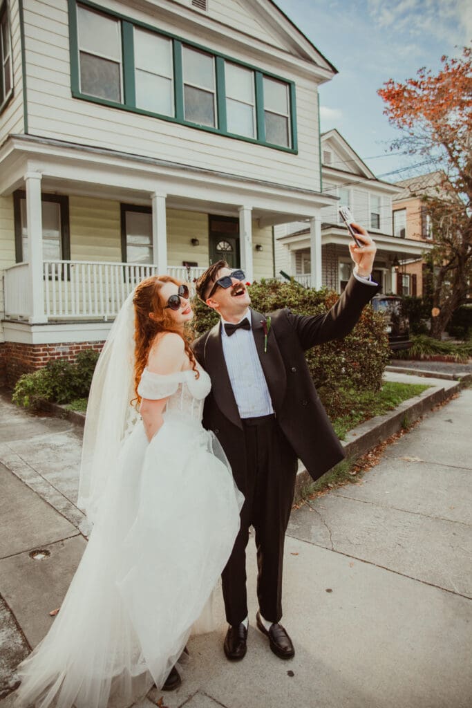 A wedding couple wearing sunglasses and taking a selfie outside a historic downtown Charleston home.