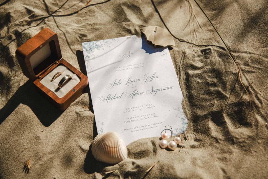 Wedding rings, an open ring box, a vow booklet, and seashells arranged on the sand for a Folly Beach elopement.