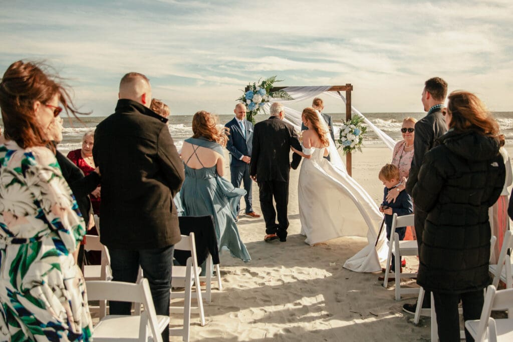 Guests watching a couple exchange vows beneath a floral wedding arch at an intimate Folly Beach ceremony near Charleston.