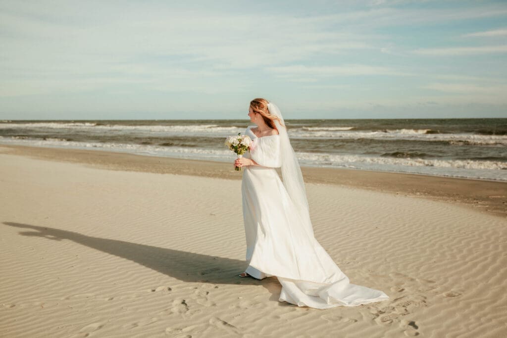 A bride looking back over her shoulder with her bouquet as her dress trails across the sand at Folly Beach.