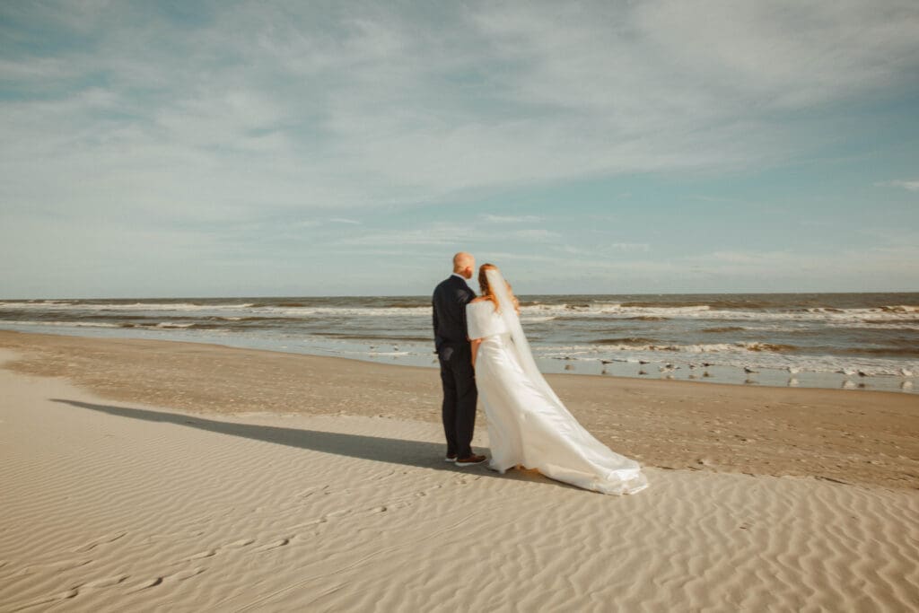 A newly married couple embracing beside the ocean as the bride’s veil blows across the sand.