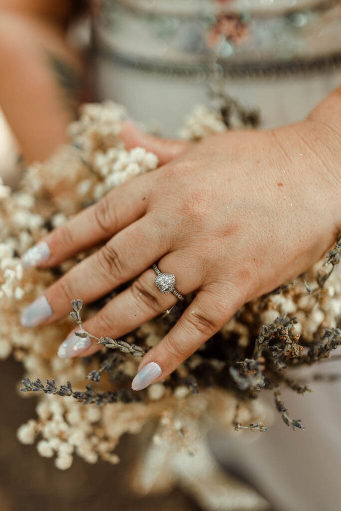 Close-up of a bride’s engagement ring and pale blue manicure resting on a dried floral bouquet.