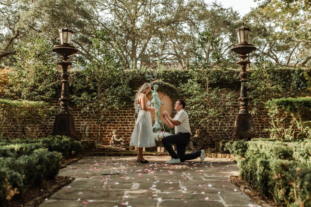 A man proposing down on one knee to a woman in green garden with rose petals on the stone path way.