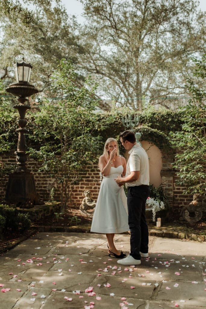 A woman holds a hand to her face as her fiancé stands beside her beneath an ivy-covered brick arch.
