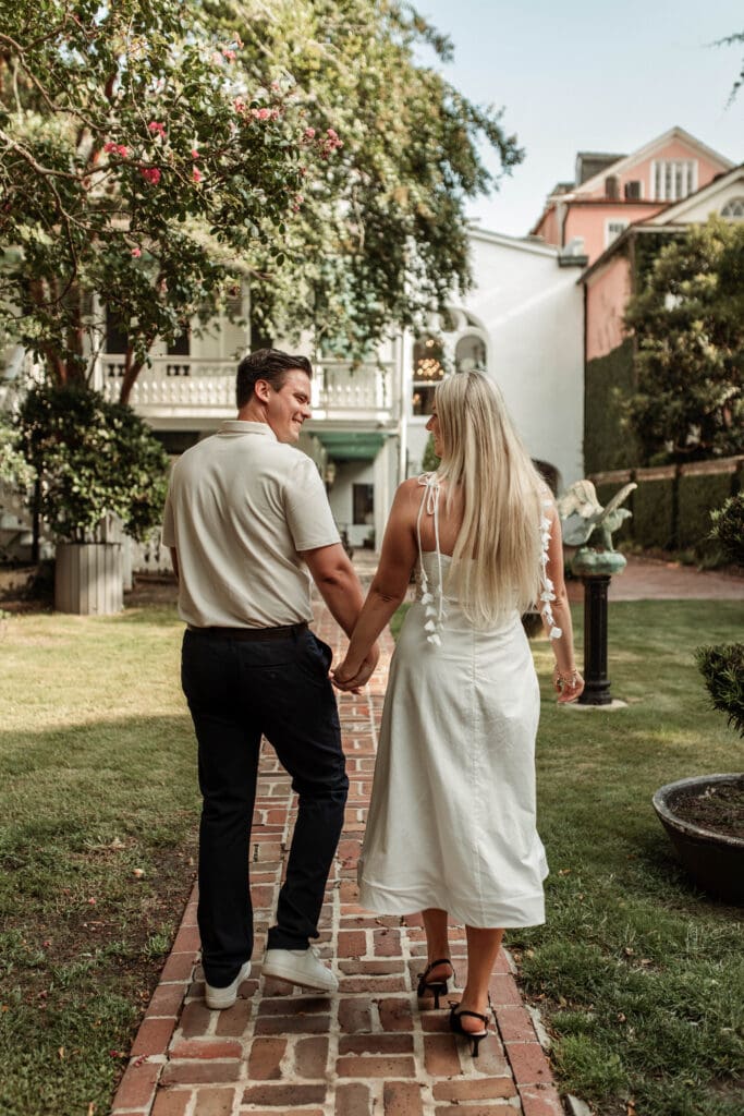 A newly engaged couple walking hand in hand along a brick garden path at 20 South Battery.