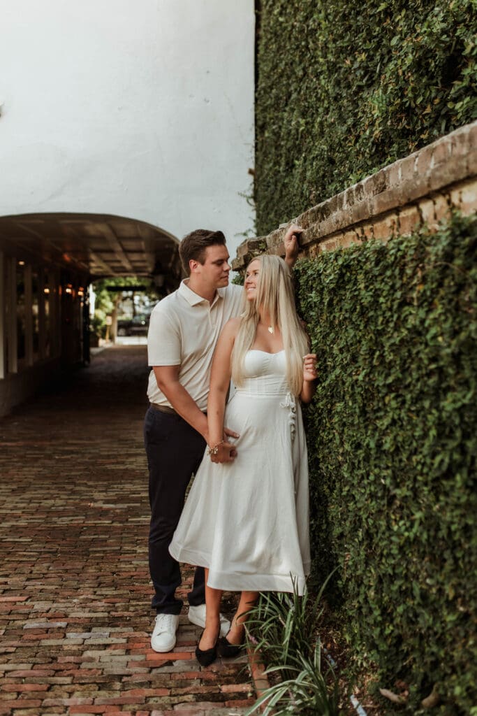 A man kissing his fiancée’s temple as they hold hands beside an ivy-covered brick wall.