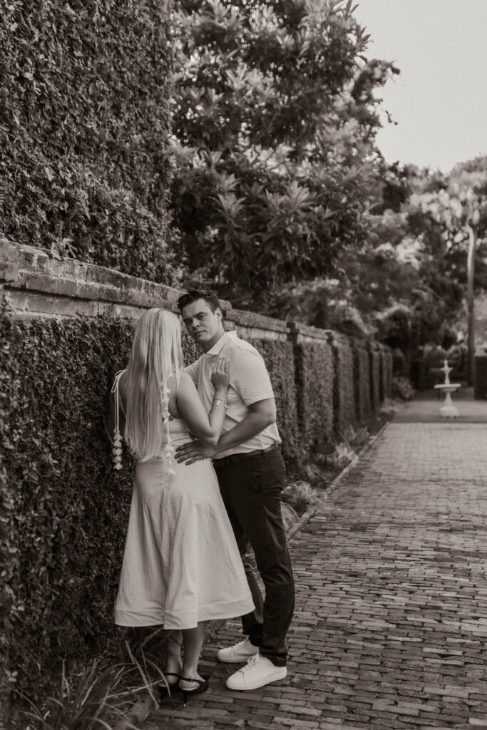 Black and white portrait of a couple embracing beside an ivy-covered brick wall.