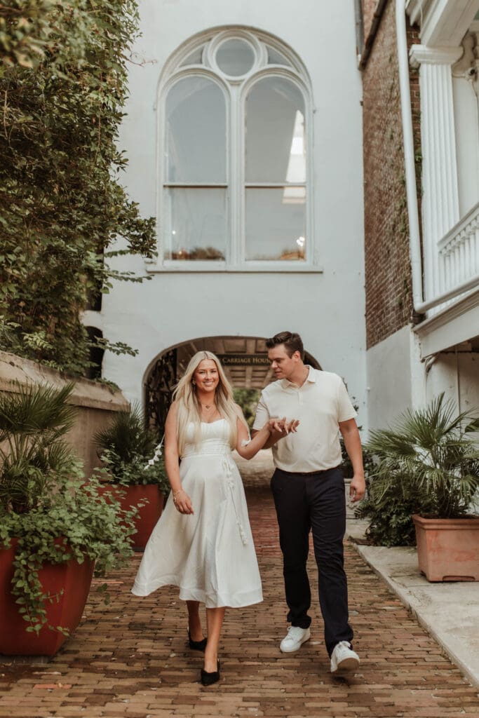 A couple walking hand in hand along a brick path beneath a tall arched window.