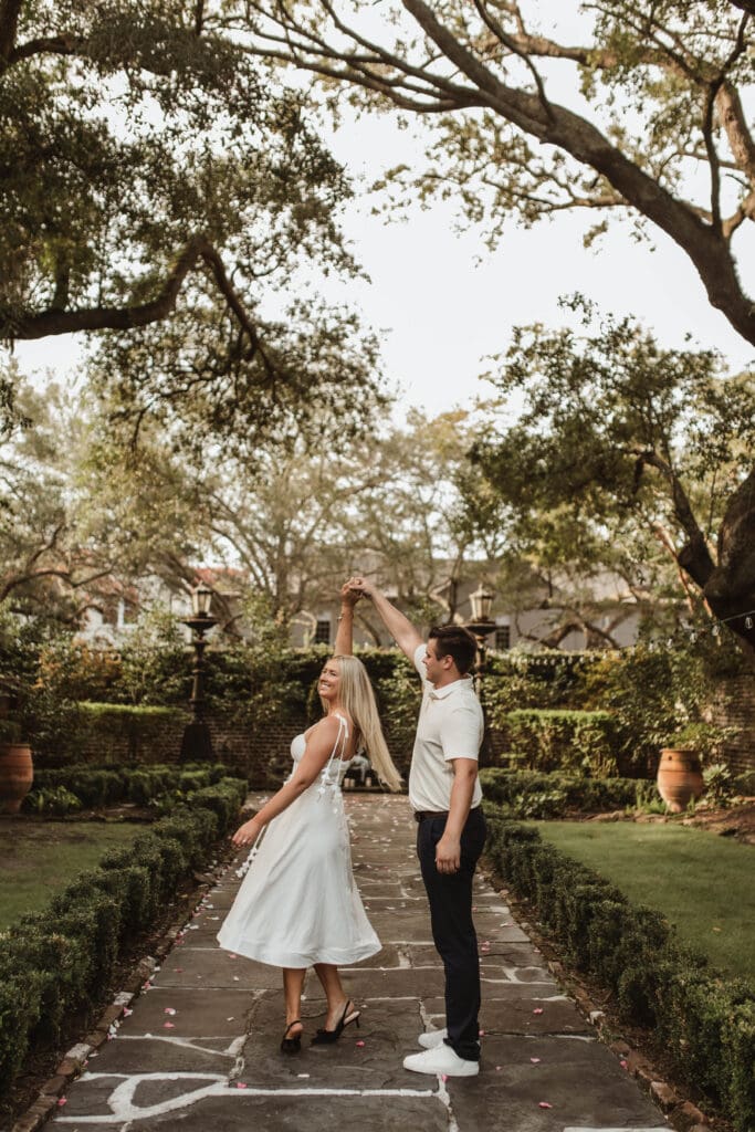 A man twirling his fiancée beneath the trees on a rose-petal-covered garden path after their Charleston proposal.