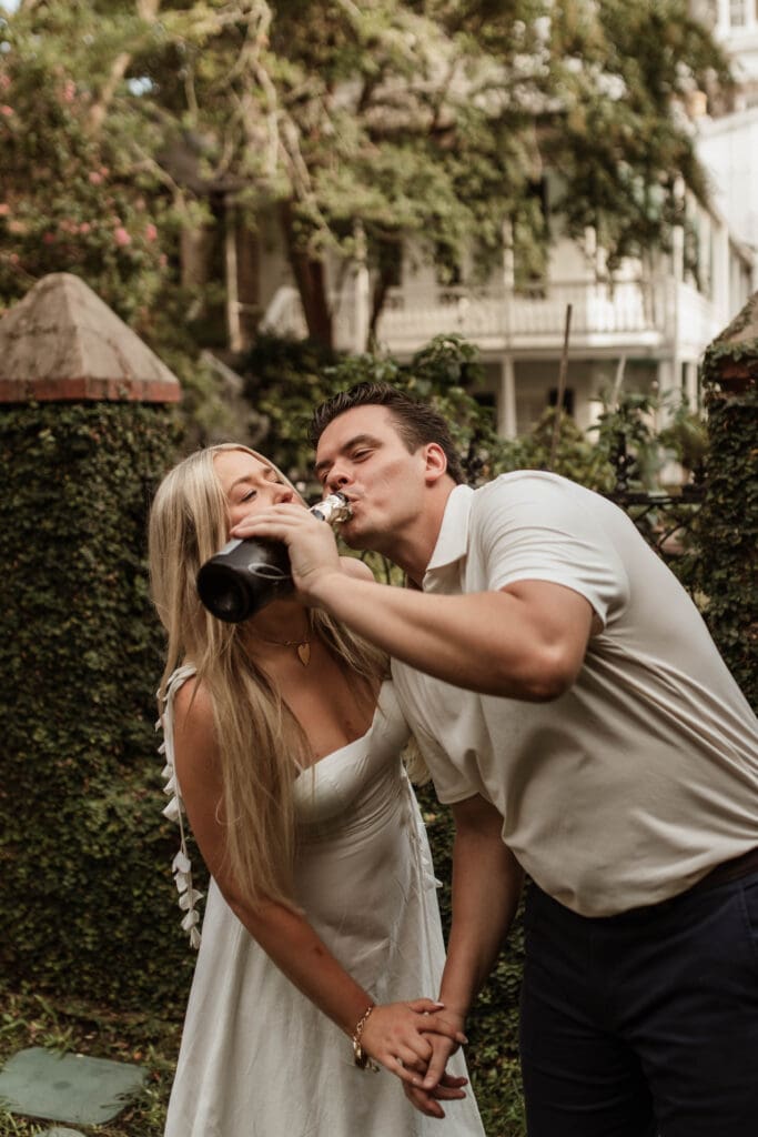 A woman laughing as her fiancé drinks champagne from the bottle while holding her hand.
