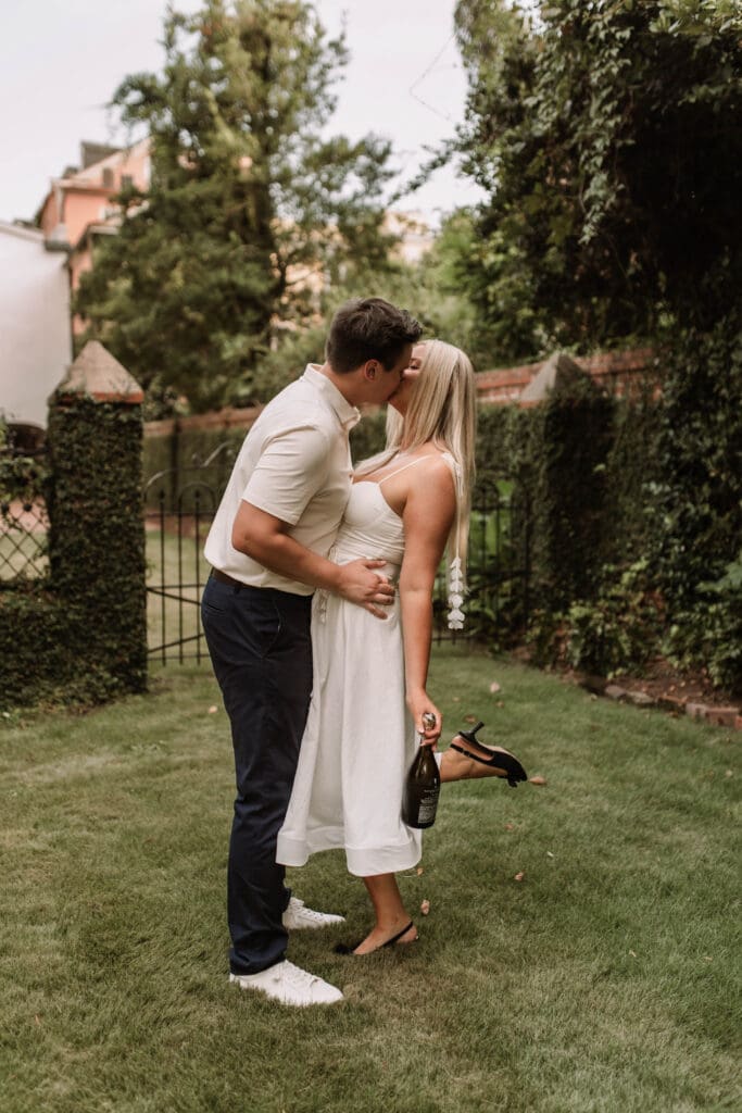 A couple kissing on the garden lawn as the woman holds her heels behind her.