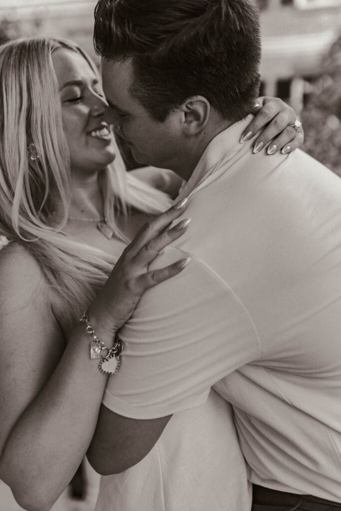 Black and white close-up of a man kissing his fiancée’s cheek as she smiles, her engagement ring visible on his shoulder.